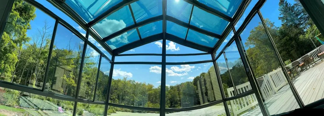 Interior view of a sunroom with a glass ceiling, featuring a ceiling fan and cozy seating, with clear blue skies and trees visible through the windows