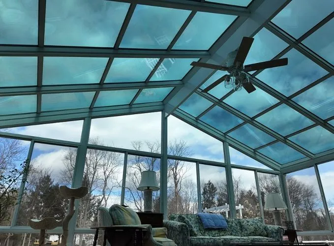 Interior view of a sunroom with a glass ceiling, featuring a ceiling fan and cozy seating, with clear blue skies and trees visible through the windows